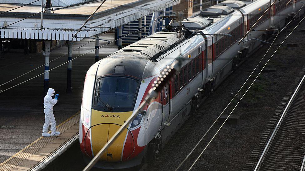 The red, yellow and grey LNER train standing at a platform in Huntingdon station. A forensic officer with white overalls stands on the platform at the front of the train. 