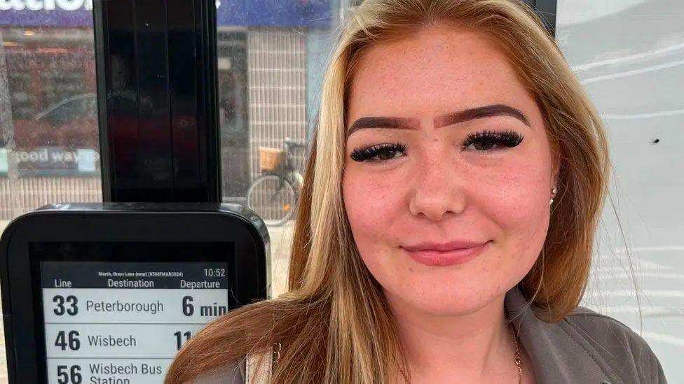 Laurisa Hallam who has long fair hair and is wearing a grey jacket. She is standing in a bus shelter and to her right is a black metal box which says the 33 to Peterborough is due to depart in six minutes.