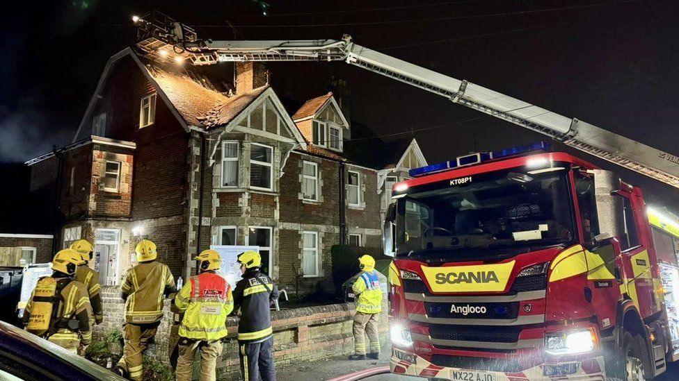 Fire engine pulled up outside a large brick house - six firefighters are standing on the pavement in front of the building - a ladder from the fire engine stretches to its roof - it is raining.