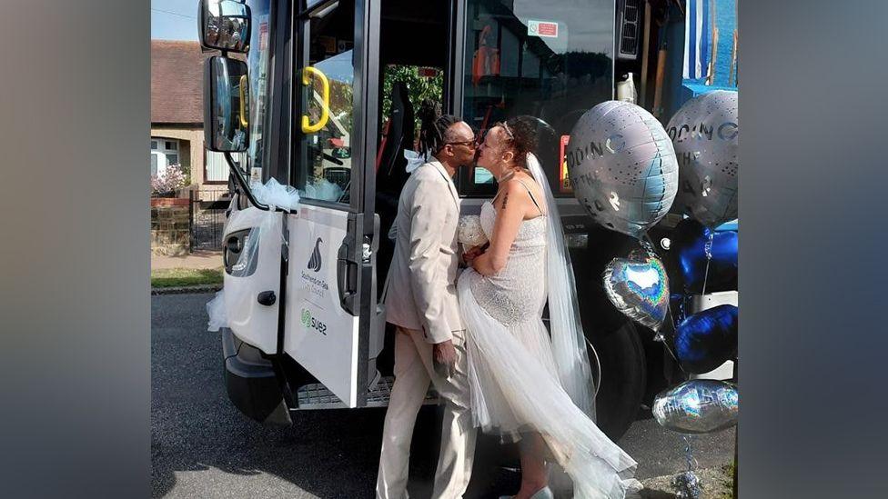 Danny and his wife are standing by the bin lorry, kissing. Balloons are next to them. The bin lorry door is open. The lorry is parked on a residential street.
