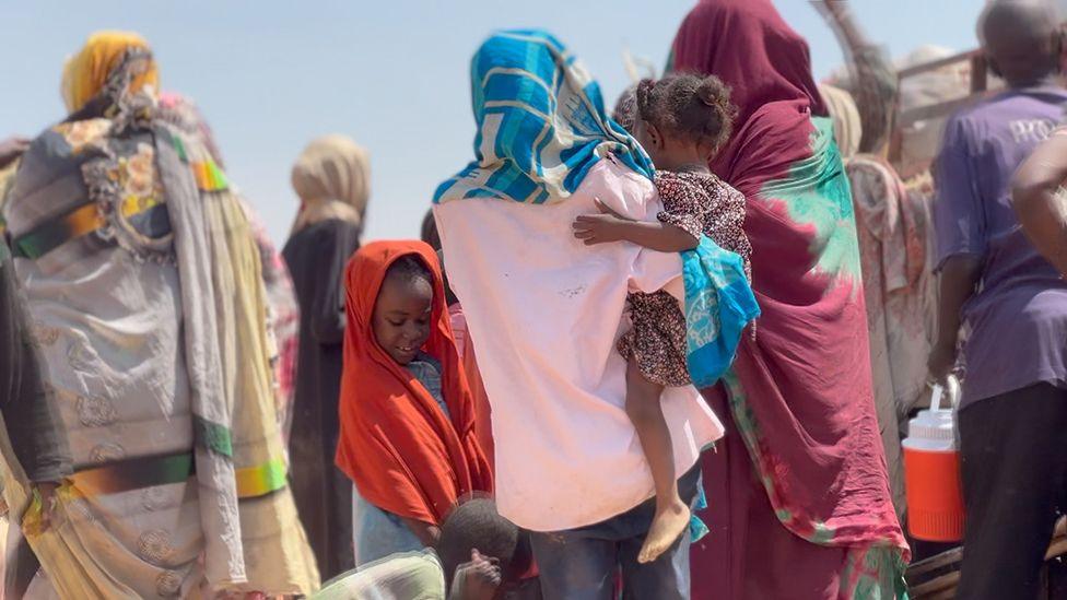 A woman, in a pink shirt and blue scarf, with her back to the camera holds a child on her hip. Another child can be seen on her left.
