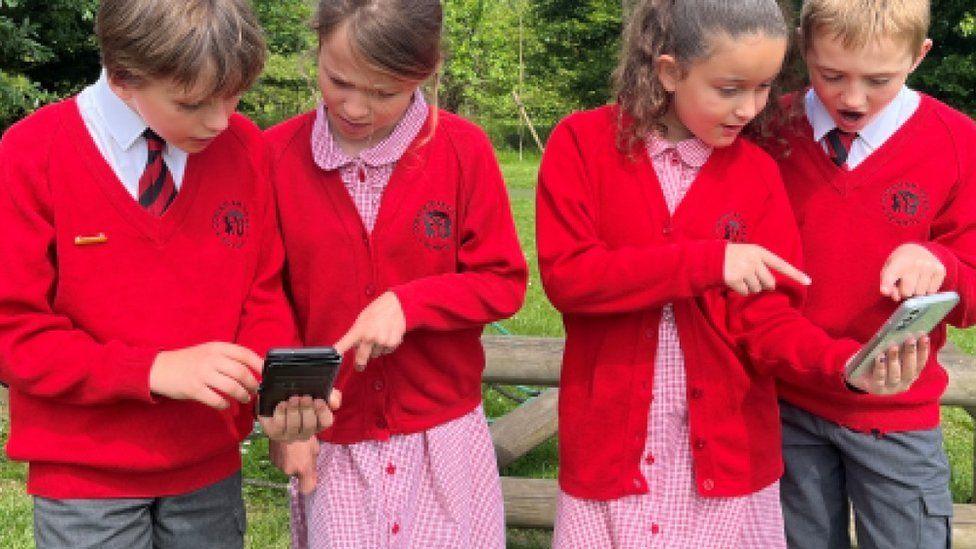 Four children in red and grey uniforms pointing at smartphones with park and trees in background