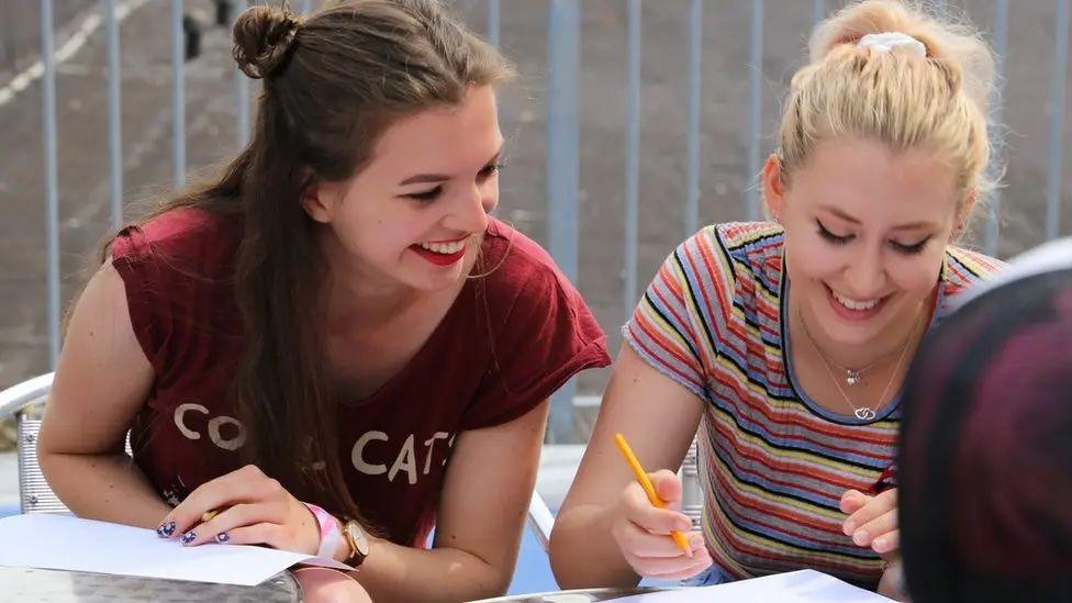 Two teenage girls wear t-shirts and smile as they look down at work. 