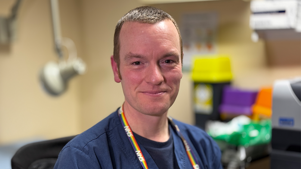 A smiling Dr Merriel wearing blue NHS scrubs and has an NHS badge around his neck in rainbow colours. He is sitting in his GP surgery and there is medical equipment in the background. 