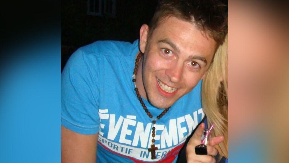 Jon Casey wearing a blue t-shirt with large white letters on it. He is smiling and wearing a necklace with a wooden cross detail hanging at its base.