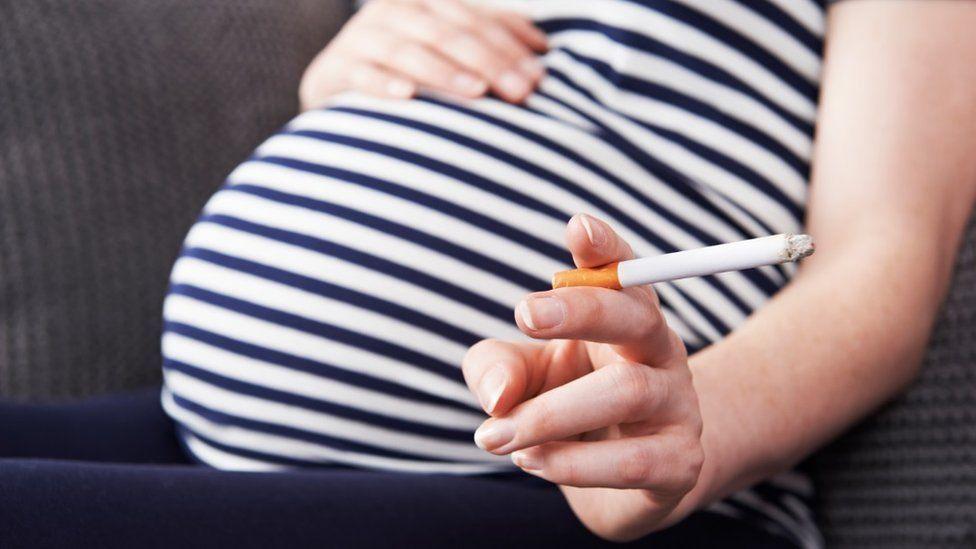 Pregnant woman with a cigarette in her left hand. You can only see her stomach and arm, she is wearing a blue and white stripped top, with blue trousers. 