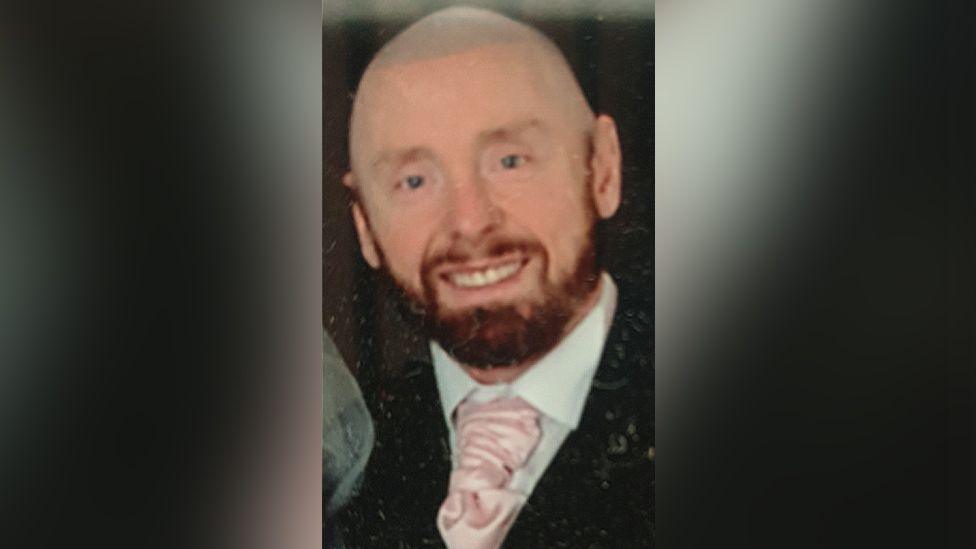 James Clacher, a bald man, with a dark beard smiling while wearing a suit and pink tie.