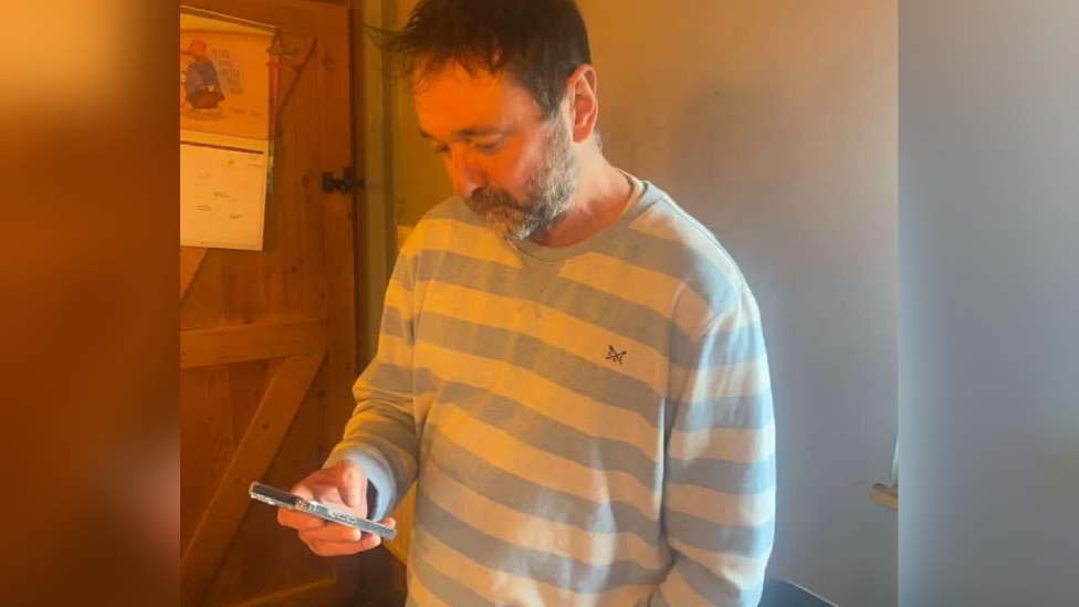A man with spiky brown hair wearing a blue and white striped jumper and looking down at a movile phone in his hand.