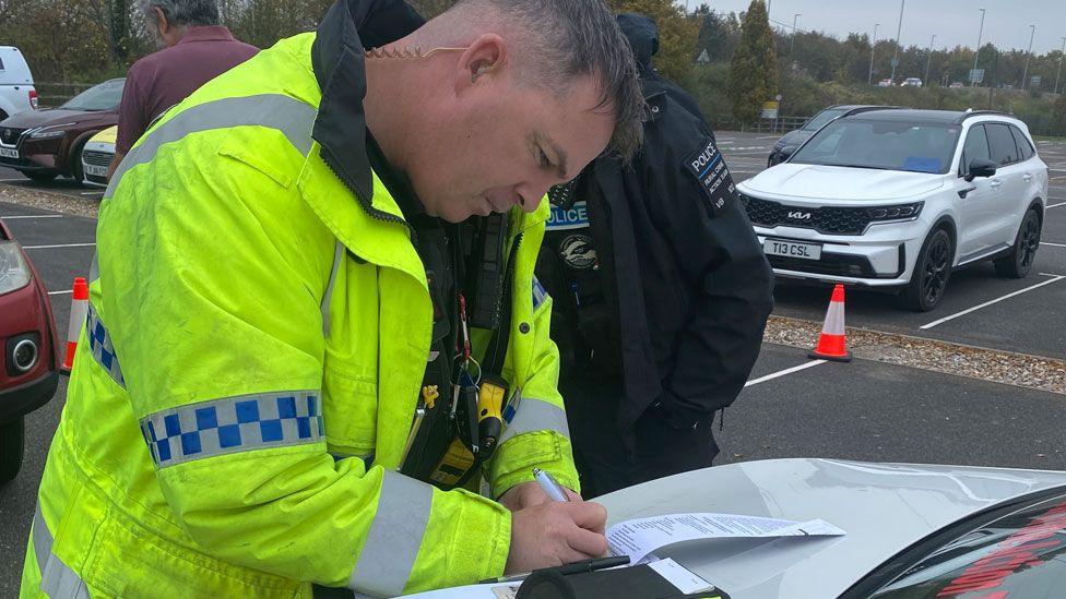 A police officer in a high-vis yellow jacket leaning over a car bonnet to write out some paperwork. Behind him can be seen more cars lined up in a car park