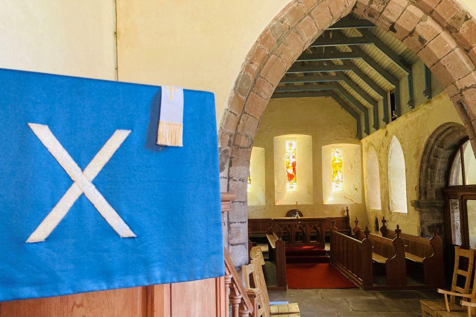 Interior of church building - Arbuthnott Church - with narrow stained glass windows in the background, and a pullpit in the foreground, with a white x on blue material draped over it.