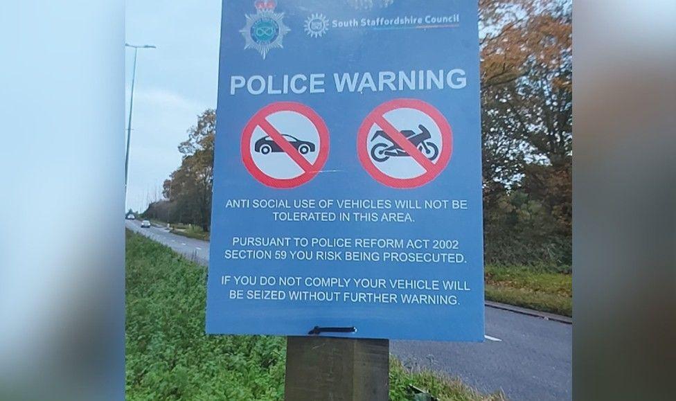 A blue rectangular road sign carrying a police warning stating anti social use of vehicles will not be tolerated. There are two circles with red borders, black symbols of a car and motorbike in each with a black diagonal line passing through them.