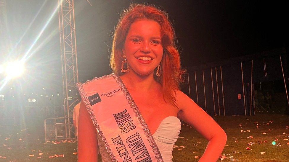 Laura Hadzopoulos wearing a white dress. She is standing underneath a big top tent looking directly at the camera and smiling. She is also wearing a Miss Universe Switzerland Finalist sash. 