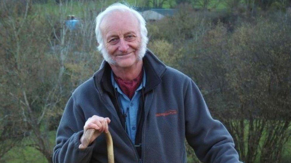 Peter Jinman smiling at the camera. He has white hair and a beard and is wearing a light blue fleece and has one hand on a stick. Behind him are trees and grass areas.