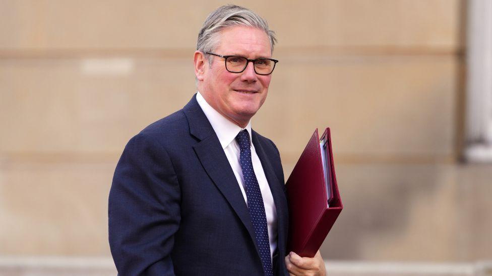 Keir Starmer, who has grey hair and glasses, carries a red folder under his left arm. He is wearing a dark blue suit and tie with a white shirt. There is a stone building in the background. 