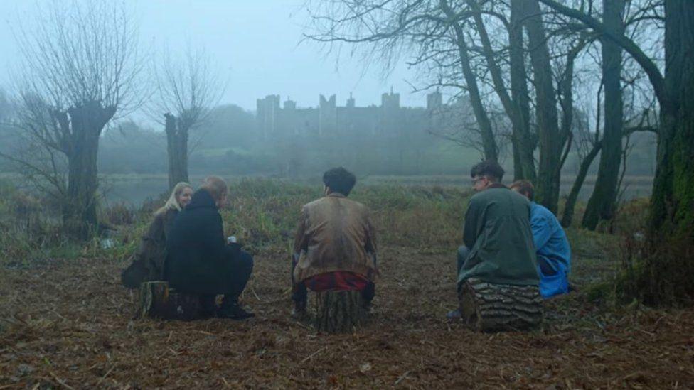 A group of people sit on wood logs in a semi circle talking in Ed Sheeran's music video for Castle on the Hill. They sit facing Framlingham Castle which is in the distance on a gloomy day.