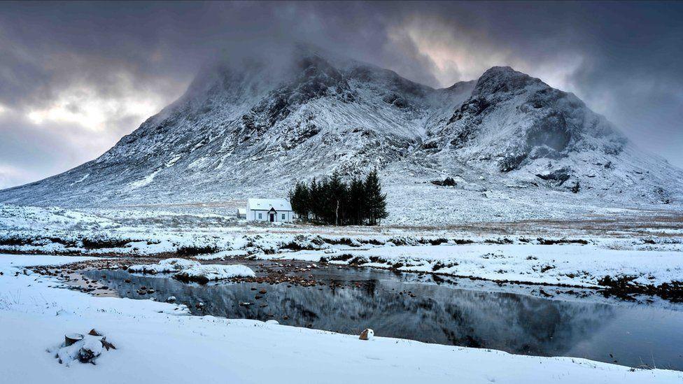 A snowy scene. There is a river in the foreground and a huge mountain in the background covered in snow. there is a tiny white house in the centre which is hard to make out due to the whole picture being white with snow.