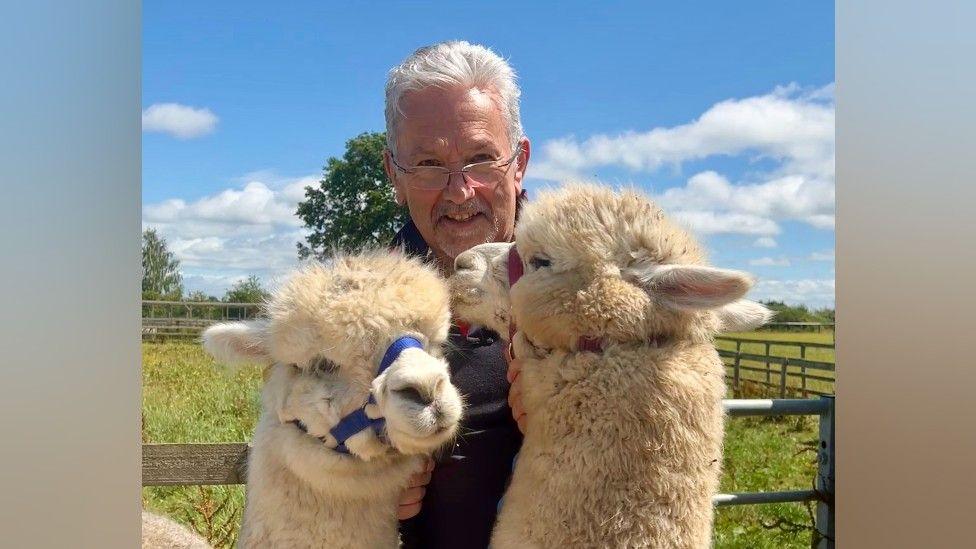 A man with white hair and glasses is posing with two fluffy cream coloured alpacas. Behind them is a wooden fence, blue sky, large tree and a field of green grass.