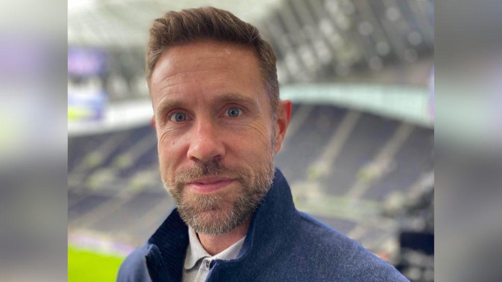 Matthew Upson, who has short brown hair and is starting at the camera with a slight smile. He has a blue quarter zip jumper on with a grey polo shirt underneath. He is standing in the Tottenham Hotspur Stadium with a large stand behind him, which is blurred in the background.