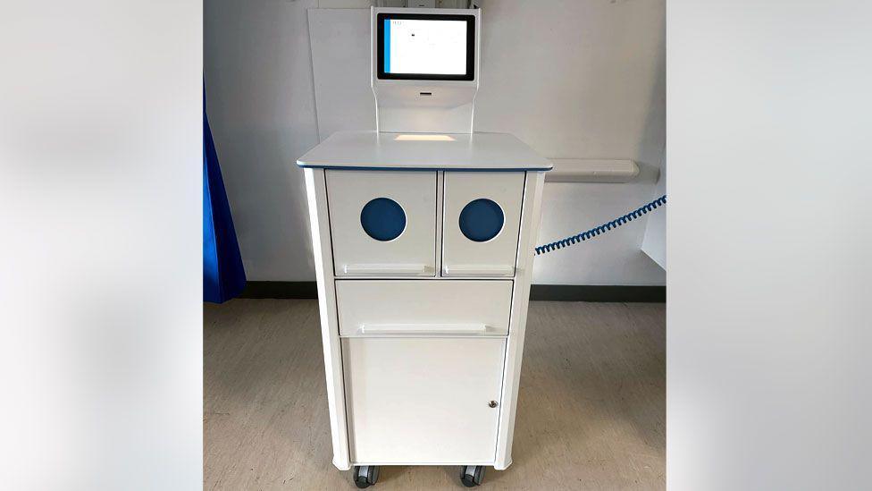 Robot trolley - white cabinet with blue markings and a screen, in a hospital room