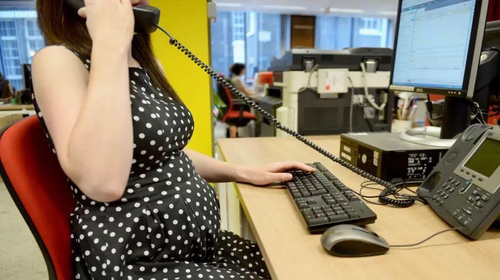 A pregnant woman in a black and white polkadot dress sitting at a desk in an office. She has one hand on the computer keyboard and the other is holding the desk phone receiver.