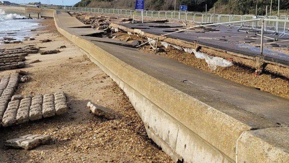 Broken up concrete seawall with twisted railings and lumps of material strewn across a beach