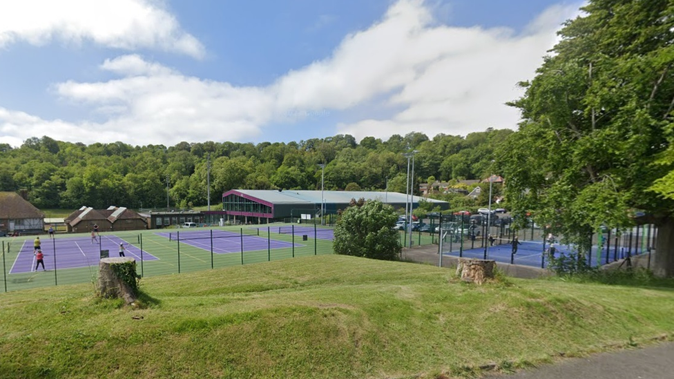 A sports complex with racquet sports courts and a pavilion. The complex is surrounded by grass and trees.