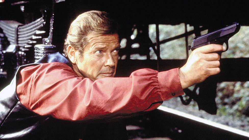 Roger Moore who has swept back light brown hair and is wearing a black leather waistcoat over a red blouson shirt. He is lying on his stomach on a railway track, with a train carriage glimpsed above him. He is stretching out his arm and a black gun is in his hand.