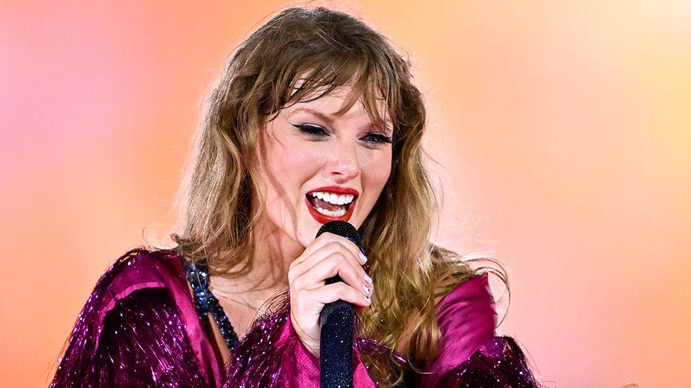 Head and shoulders shot of Taylor Swift singing into a microphone while wearing a purple dress against a peach coloured background, at Wmbley in London on 15 August