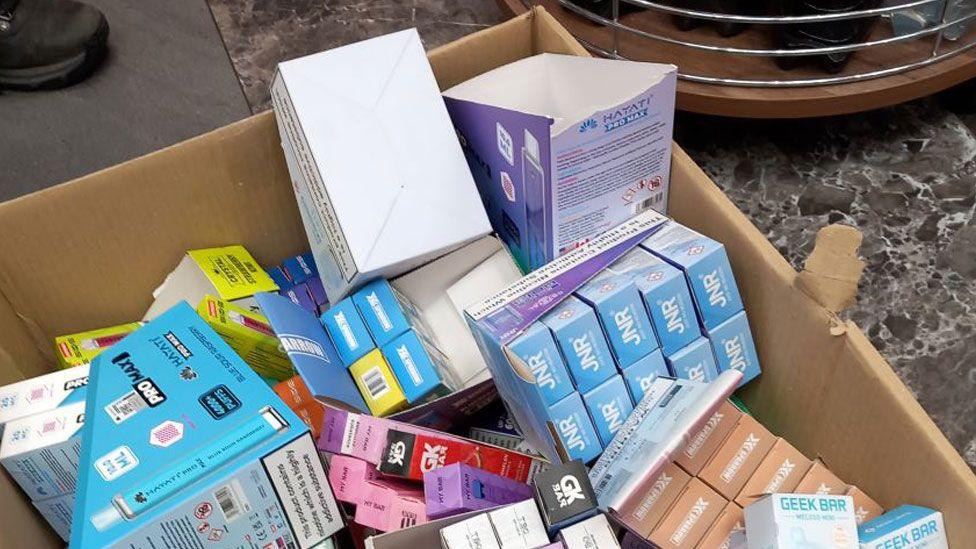 A large brown cardboard box, resting on a grey floor, filled with smaller colourful branded boxes.