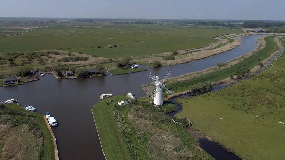 A bird's eye view of the Broads with several small boats on the water. A windmill is on the field next to the water