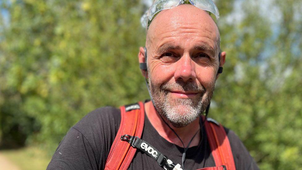 Gavin Ward who has a short salt and pepper beard and has clear goggles on the top of his head. He is wearing a black T-shirt and over it are red straps. Behind him are blurred out trees and grass.