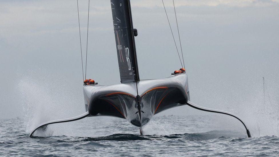 High-tech design racing yacht with foils in the water on both sides head with orange helmets seen either side of the boat with black sail coming up the centre