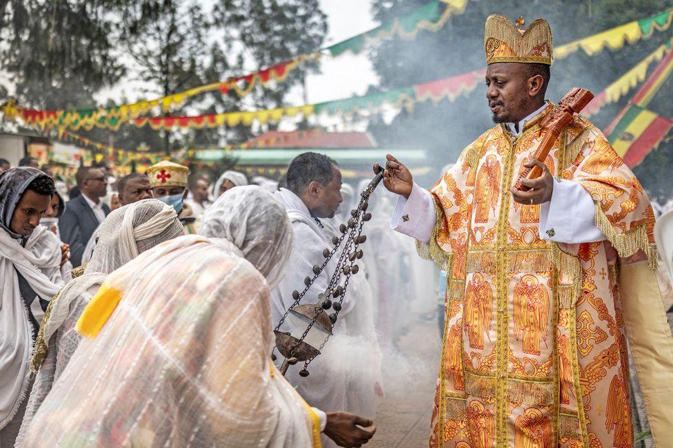 An Orthodox priest in ornate orange and gold robes swings an incense burner towards people dressed in white scarves and shawls. They bow towards him at Entoto St Raguel Church in Addis Ababa, Ethiopia - Thursday 11 September 2025.