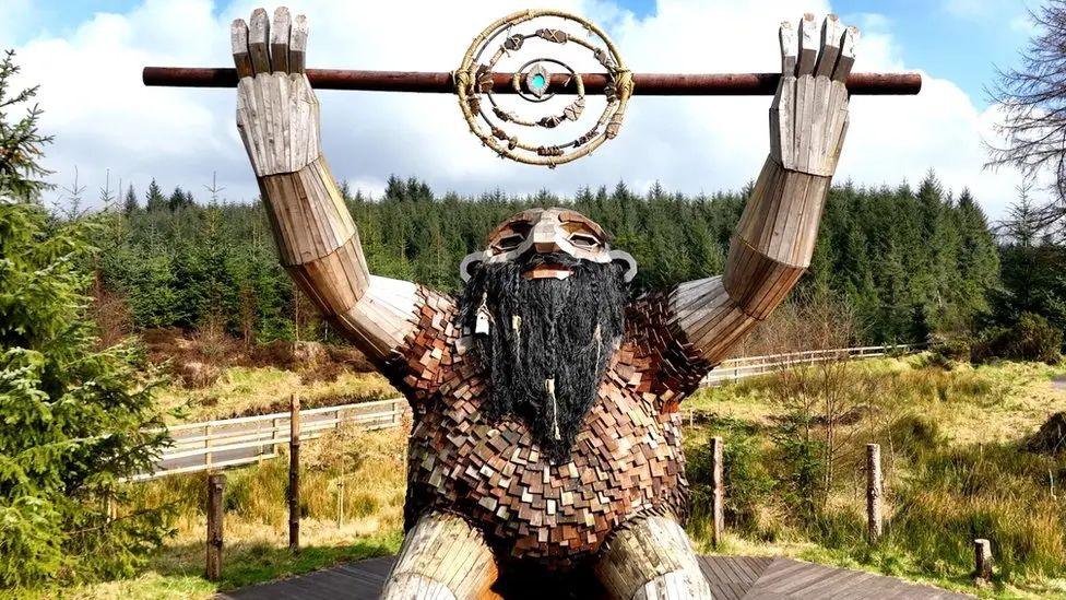 A troll made of wood holds a stick in the air. There is a spiral in the middle with a glowing centre.