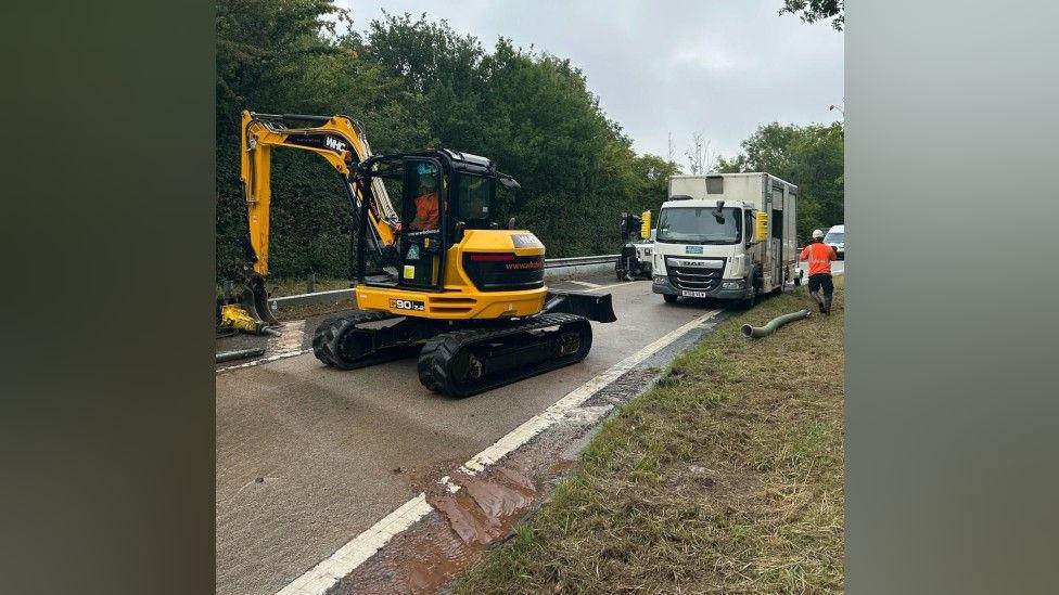 A yellow digger is working on a slip road that has brown mud smeared over it, as well as some standing water. There is a large white Severn Trent lorry in the background with a workman in orange hi-vis clothing.