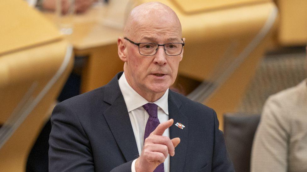 John Swinney, who is bald with glasses, speaks with his right hand raised in front of him, with his index finger pointing ahead. He is wearing a dark suit, white shirt and purple tie.