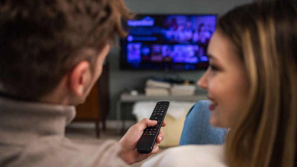 Rear view of cheerful couple smiling and talking while choosing a movie on TV. 