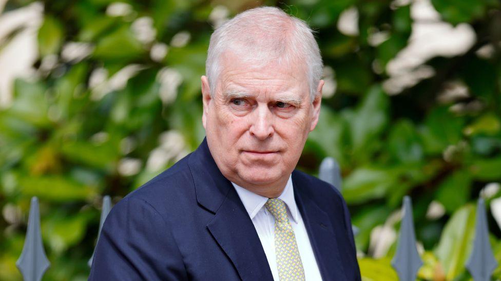 Andrew at  St George's Chapel, Windsor Castle in April 2025. He's wearing a green patterned grey tie and navy suit. Behind him are trees and a metal fence