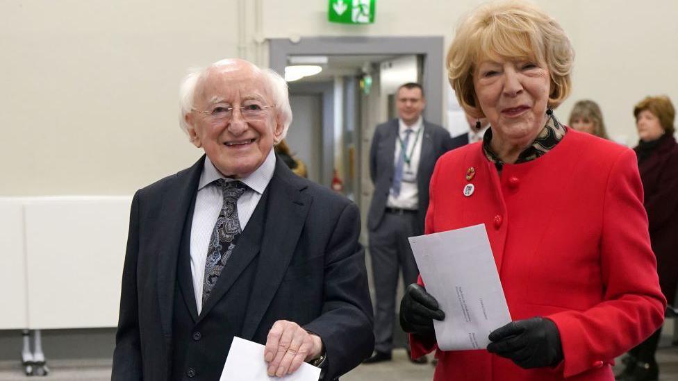 Michael D Higgins in black suit and white shirt, wife Sabina in red coat and black gloves, holding ballot papers and voting