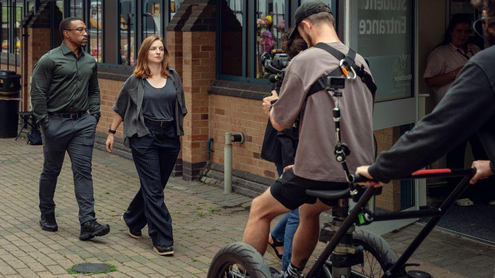 Ashley Walters and Erin Doherty walking down a street while being filmed by a cameraman ahead of them
