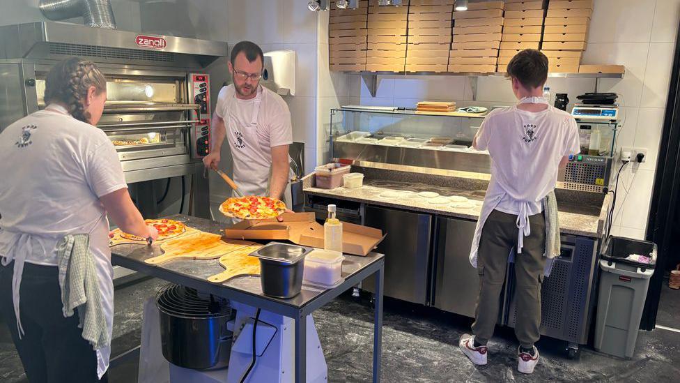 Three people in a kitchen making pizzas, one at a prep counter, one putting pizzas in the oven and the third one slicing them