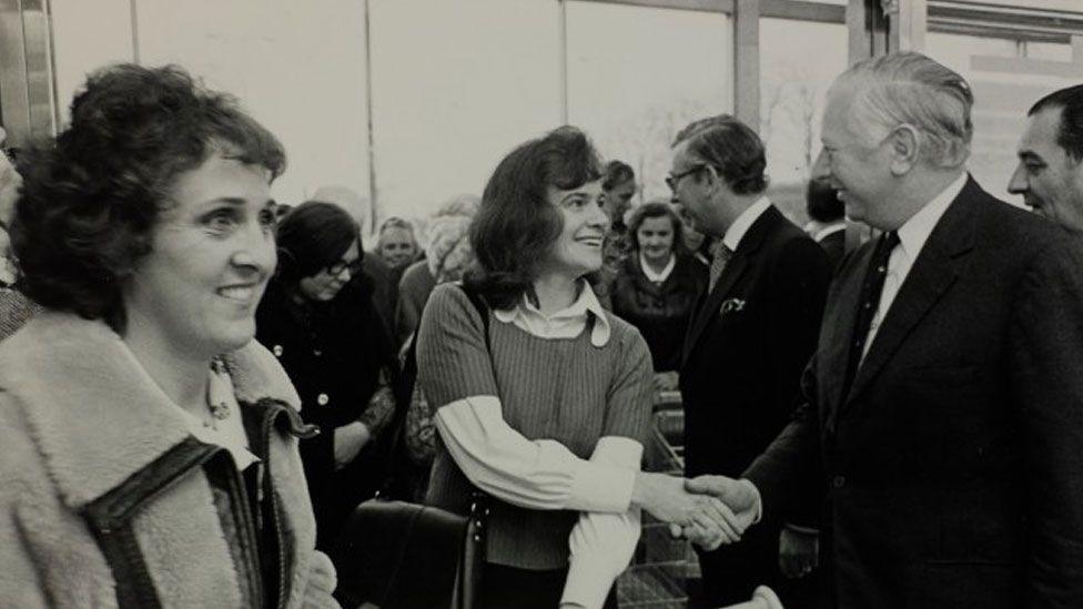 A black and white image showing women coming in through the entrance of a supermarket in 1975. One in the middle has shoulder-length hair and is wearing a short sleeved jumper over a white shirt and  is shaking hands with MP Jim Prior. He is wearing a dark suit and tie and has slicked-back grey hair