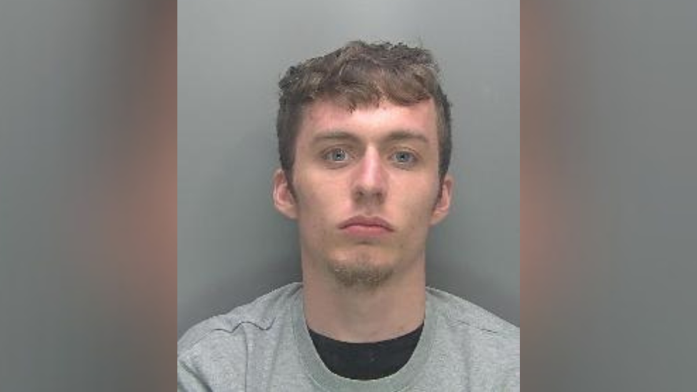 Custody photo of Callum Schooling against a plain grey background. He is wearing a crew neck grey sweatshirt over a crew neck black t-shirt. He looks directly at the camera and is not smiling. He has brown hair, blue eyes and facial hair on his chin.