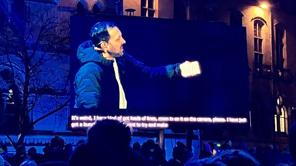 Stephen Frayne shown on a big screen holding up a card