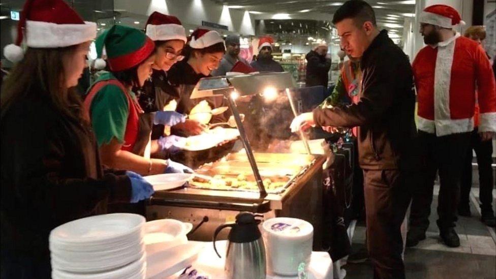 A group of people queue up to take food from a hot food counter inside a building. The people on both sides of the counter are wearing red and white Santa hats. The building has a grey ceiling with several white spotlights.