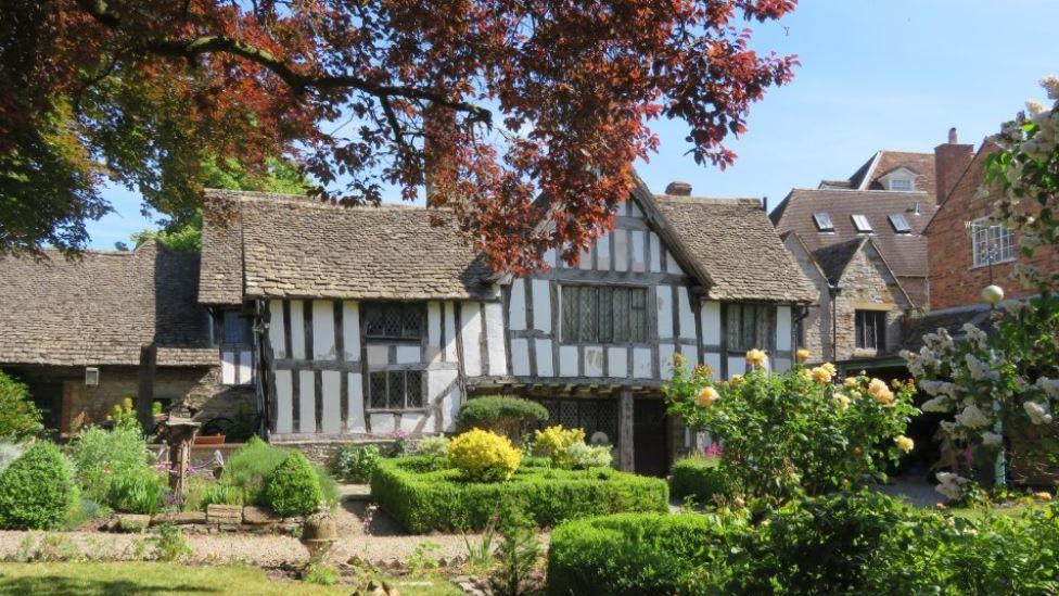 The front of a large black and white timbered building with a green garden in front of it and a tree with red leaves overhanging it