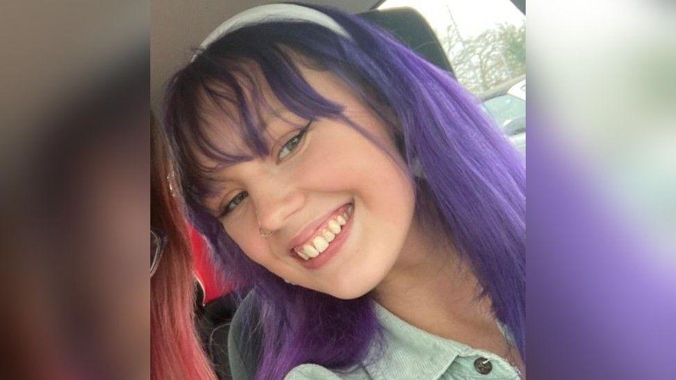 A close up photo of Evelyn with long purple hair, a fringe and she is smiling to the side into the camera and wearing a denim shirt.