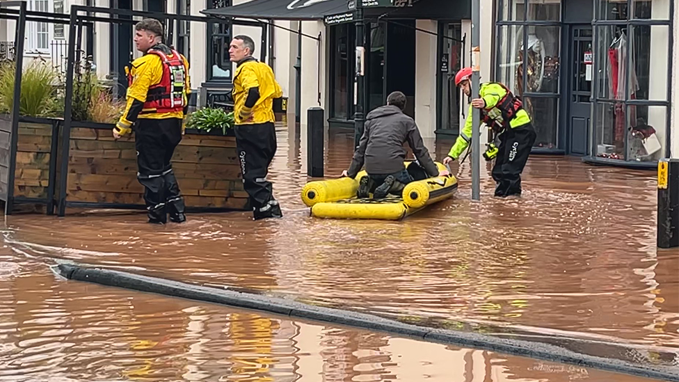 A man sits in a small dinghy as he is ferried by a rescuer wading through water