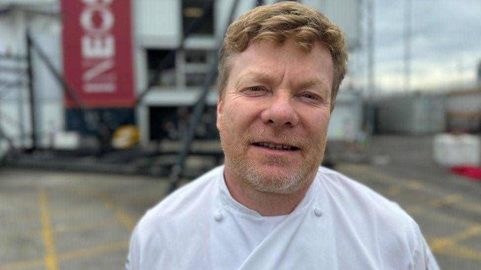 Man with short blonde hair with stubble wearing chef whites standing infron of a building with red and white Ineos sign