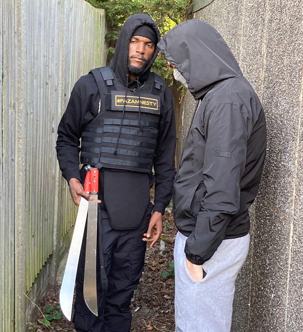 Faron Paul who runs a knife amnesty company is standing in an alleyway holding two large knives which were handed to him by Jon (not his real name) who is standing next to him wearing a hood and a covering over his mouth.